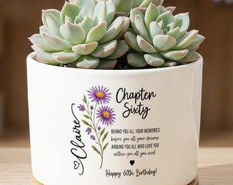 Op de afbeelding: Een witte keramische plantenpot met een bamboe onderzetter en een groene vetplant. De pot is voorzien van een paarse bloemenillustratie en de tekst: Claire, Chapter Sixty, Behind you all your memories, before you all your dreams, around you all who love you, within you all you need, Happy 60th Birthday.