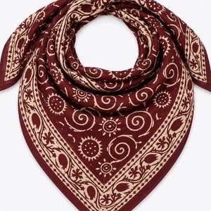 Könnte beinhalten: Ein quadratisches Bandana mit tiefburgunderrotem Hintergrund und einem kontrastierenden cremefarbenen Blockdruckmuster. Das Design zeigt stilisierte Sonnen, wirbelnde Spiralen und florale Motive, die in einem zentralen Feld und einem breiten Rand angeordnet sind.