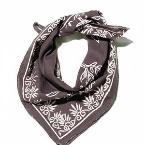 Può includere: Una bandana grigio scuro con un motivo floreale e a volute bianco. La sciarpa quadrata è piegata e annodata, mostrando il design dettagliato sui bordi. Il tessuto appare morbido e leggero, adatto a vari usi.