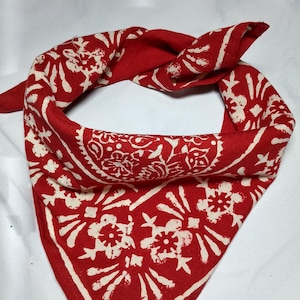 Op de afbeelding: Rode bandana met een wit bloemenpatroon. De vierkante sjaal is gevouwen en geknoopt, wat het ingewikkelde ontwerp laat zien. De stof is helderrood met een herhalend wit bloemmotief, wat een klassiek en veelzijdig accessoire creëert.