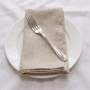 May include: A folded beige linen napkin and a silver fork rest on a white plate. The plate sits on a white tablecloth. The fork is positioned diagonally across the napkin.