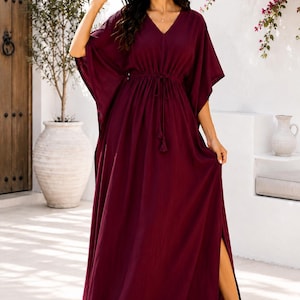May include: A floor-length, deep burgundy kaftan dress featuring a V-neckline, wide kimono-style sleeves, and a drawstring waist with tassel ties. The garment includes a high side slit along the skirt, providing a loose, flowing silhouette.