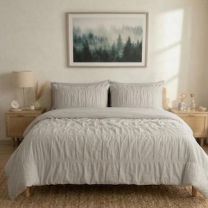 May include: A light grey bedspread and pillow set on a bed with a framed forest landscape art piece above. The bedroom has two wooden bedside tables with lamps and decorative items.