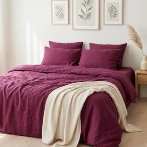 Könnte beinhalten: Ein Schlafzimmer mit einem Bett, das mit einer tiefroten Bettdecke und Kissen ausgestattet ist. Eine cremefarbene Decke ist über dem Fußende des Bettes drapiert. Drei gerahmte botanische Drucke hängen über dem Bett und verleihen dem Raum eine natürliche Note.