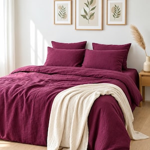 May include: A deep magenta bedding set featuring a duvet cover and four matching pillowcases. A textured cream-colored throw blanket is draped across the foot of the bed. The bedding has a visible linen-like weave and a solid, rich color.
