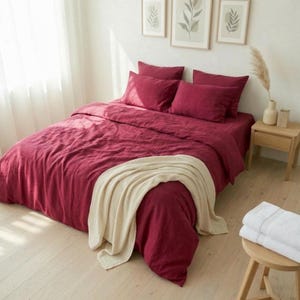 May include: A bed dressed in a deep red duvet cover and pillows. A cream-coloured throw blanket is draped over the foot of the bed. The room has a light, airy feel with framed botanical prints on the wall.