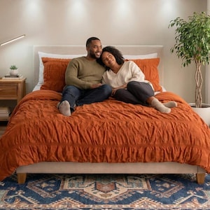 Op de afbeelding: Een slaapkamer met een oranje dekbedovertrek en bijpassende kussens op het bed. Een stel zit op het bed. Een houten nachtkastje met een lamp en een kleine plant staat links. Een patroonkleed ligt op de vloer.