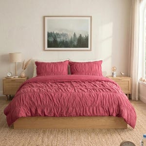 Puede incluir: Una habitación con una cama vestida con un edredón y almohadas de color rosa-rojo. La cama está sobre un marco de madera, con una obra de arte enmarcada de un paisaje forestal encima. Mesitas de noche de madera con lámparas a ambos lados de la cama.