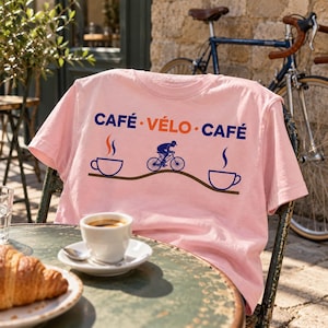Könnte beinhalten: Ein hellrosa T-Shirt mit einem Grafikdruck, der den Text CAFÉ VÉLO CAFÉ in Blau und Orange zeigt. Das Design umfasst zwei blaue Kaffeetassen-Symbole mit Dampf und eine zentrale blaue Silhouette einer Person auf einem Fahrrad.