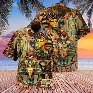 May include: Short-sleeved button-down shirt featuring an all-over print of ancient Egyptian pharaohs, hieroglyphs, and symbols in shades of gold, blue, and brown. The shirt has a classic collar and is set against a tropical beach scene with palm trees and a sunset.