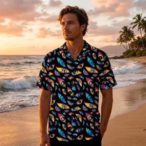 May include: Black button-down shirt with a vibrant all-over eye pattern in shades of blue, yellow, pink, and green. The short-sleeved shirt has a classic collar and is worn on a beach with a sunset backdrop.