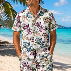 Könnte beinhalten: Ein Set aus einem kurzärmeligen Hemd mit Knopfleiste und passender Shorts auf weißem Grund mit einem wiederkehrenden Muster aus bunten Fahrrädern, grünen Palmblättern und hellrosa Hibiskusblüten. Das Hemd hat einen Kragen.
