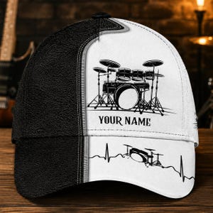 Op de afbeelding: Een baseballpet met een gespleten ontwerp met een getextureerde zwarte kant en een witte kant. Het witte gedeelte toont een zwarte afbeelding van een drumstel, de tekst "YOUR NAME" in een schreeflettertype en een hartslaglijn met een drumstel.
