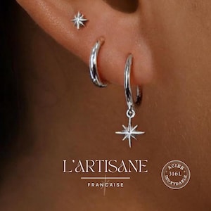 以下が含まれることがあります： 3つのシルバーのジュエリーが飾られた耳のクローズアップ。小さな星型のスタッドピアス、小さなフープピアス、そして星のチャームが揺れるフープピアス。ブランド名「L'ARTISANE FRANCAISE」が下に表示されています。