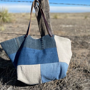 May include: A patchwork tote bag made from denim and beige fabric, with brown leather handles. The bag is hanging on a wooden post with a barbed wire fence in the background. The bag has a rectangular shape.