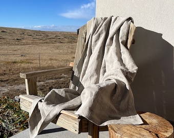 Könnte beinhalten: Eine hellbeige Leinen-Wurfdecke, die über einem hölzernen Adirondack-Stuhl drapiert ist. Der Stuhl steht auf einer Beton-Terrasse neben einem Baumstumpf. Der Hintergrund zeigt ein Feld und einen klaren blauen Himmel.