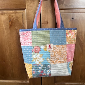 May include: A patchwork tote bag with a variety of floral and geometric patterns in shades of blue, pink, orange, and green. The bag has blue handles with a pink lining and a small tag that reads "Ruby Raindrop".