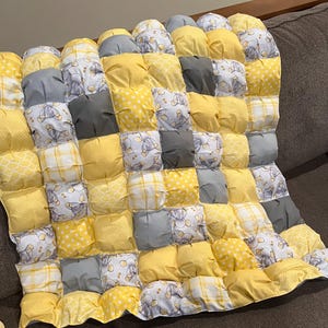 Op de afbeelding: Een handgemaakte babyquilt met een patchwork-ontwerp. De quilt heeft een patroon van gele, grijze en witte vierkanten. Sommige vierkanten hebben een stippenpatroon, andere hebben een ruitpatroon en sommige hebben een patroon van olifanten.