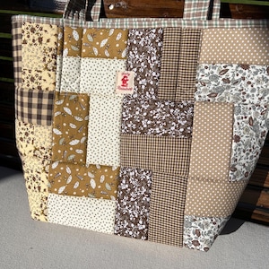May include: A quilted tote bag featuring a patchwork design of various brown, beige, and white fabrics with floral, polka dot, and gingham patterns. A small rectangular label with the text Ruby Raindrop is stitched onto the front center.