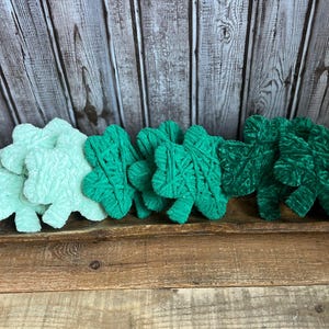 May include: A collection of plush shamrock decorations in varying shades of green and a light teal color. The shamrocks are arranged in a wooden bowl, perfect for St. Patrick's Day or other festive occasions.