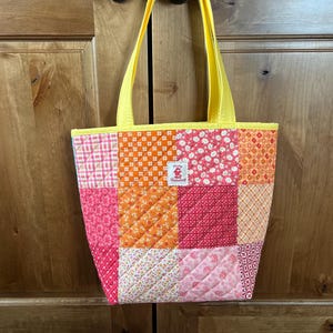 Puede incluir: Bolso tote acolchado con un diseño de patchwork en tonos rosa, naranja y rojo. El bolso tiene asas y ribetes amarillos. Una pequeña etiqueta con texto es visible.