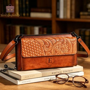 May include: A tan leather crossbody bag featuring a textured flap with a woven pattern and floral embossing. The bag has a smooth lower section with the letter E printed in the centre, a black zip closure, and a matching adjustable shoulder strap.