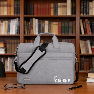 May include: A gray laptop bag with a black shoulder strap and handle. The bag has the name "Brooke" printed on the front in white script. A pair of glasses and a pen are on the table in front of the bag.