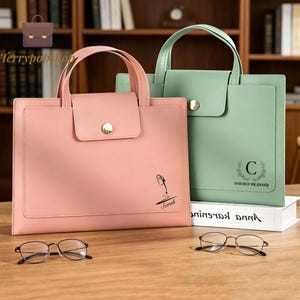 May include: A pink document bag with a pen graphic and the name Sarah, and a green document bag with a letter C and floral wreath graphic. Both bags feature top handles, a flap closure with a gold button, and are paired with two pairs of glasses.