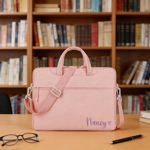 May include: A pale pink laptop bag with a shoulder strap and a top handle. The bag has a zip closure and the name "Nancy" is written in cursive with a heart symbol. The bag is on a wooden desk.