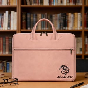 Può includere: Borsa per laptop rosa cipria con manico superiore e cerniera frontale. La borsa presenta una grafica di drago nero e il nome "MASON" stampato sotto. La borsa è appoggiata su una superficie di legno davanti a una libreria.