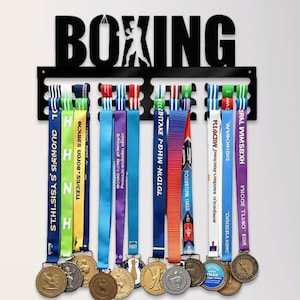 May include: A black metal boxing medal display rack with the word "BOXING" and a boxer silhouette. Colourful ribbons with medals hang from the rack. A collection of gold trophies sits below.
