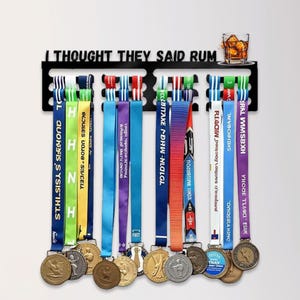 May include: A black metal medal display rack with the text "I THOUGHT THEY SAID RUM" and a small glass of liquor. Below, a collection of gold trophies and a variety of colorful medals.