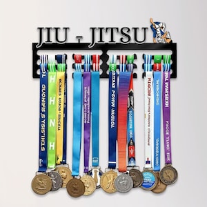 Op de afbeelding: Een zwartmetalen Jiu-Jitsu medaille display met kleurrijke linten en medailles. Daaronder staat een verzameling gouden en zilveren trofeeën van verschillende formaten op een tafel.
