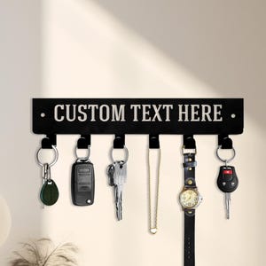 May include: A black key holder with the text "CUSTOM TEXT HERE" in white. The key holder has five hooks, each holding different items, including keys, a car remote, a necklace, and a watch. The key holder is mounted on a white wall.