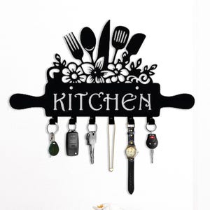 May include: Black metal kitchen key holder with a rolling pin design. The word "KITCHEN" is displayed in white letters. The key holder features a floral and utensil design at the top and has hooks for keys and other items.