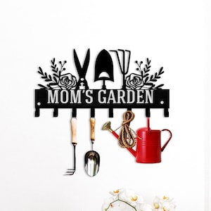 May include: Black metal garden tool organizer with the text "MOM'S GARDEN". The organizer features a variety of garden tools, including a rake, trowel, and watering can. The watering can is red, and the other tools have wooden handles.