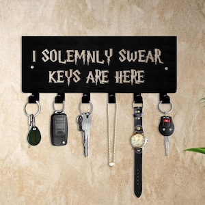 以下が含まれることがあります： 「I SOLEMNLY SWEAR KEYS ARE HERE」の文字がスタイリッシュなフォントで書かれた黒い木製のキーホルダー。キーホルダーには、さまざまな鍵、時計、ネックレスがぶら下がっている6つのフックがあります。キーホルダーはベージュ色の壁に取り付けられています。