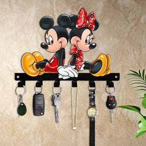 May include: Black metal key holder featuring Mickey and Minnie Mouse. The key holder has six hooks with keys, a watch, and a necklace hanging from them. Mickey Mouse is wearing red shorts and a red shirt. Minnie Mouse is wearing a red dress with white polka dots.