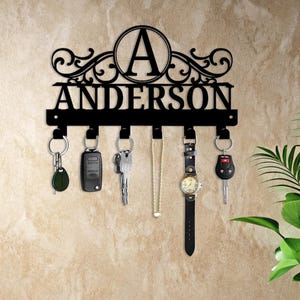 May include: Black metal key holder with the name "ANDERSON" and the letter "A" in a circle. The key holder has hooks with keys, a car key fob, a necklace, and a watch hanging from it. The key holder is mounted on a beige wall.