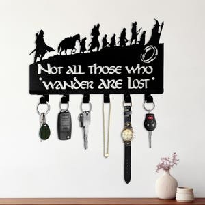 May include: Black metal key holder with a silhouette of figures and the text "Not all those who wander are lost." The key holder has six hooks with keys, a watch, and a necklace hanging from them. A small vase with flowers sits on a wooden table.