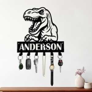 May include: Black metal key holder featuring a Tyrannosaurus Rex design with the name "ANDERSON" below. The key holder has multiple hooks with keys, a watch, and a necklace hanging from them. The key holder is mounted on a white wall.