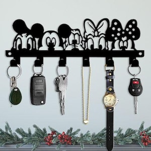 May include: Black metal key holder featuring Mickey Mouse, Minnie Mouse, and friends. The key rack holds keys, a watch, and a necklace. The key holder is mounted on a white wall above a gray dresser.