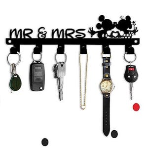 Könnte beinhalten: Schwarzer Metall-Schlüsselhalter mit den Worten "MR & MRS" und Mickey Mouse-Silhouetten. Der Schlüsselhalter hat sechs Haken mit Schlüsseln, einer Uhr und einer Halskette. Der Hintergrund ist weiß mit roten, gelben und schwarzen Mickey Mouse-Formen.