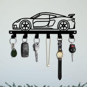 May include: Black metal key holder shaped like a sports car, with five hooks holding keys, a car key fob, a watch, and a necklace. The key holder is mounted on a white wall above a gray dresser decorated with greenery.
