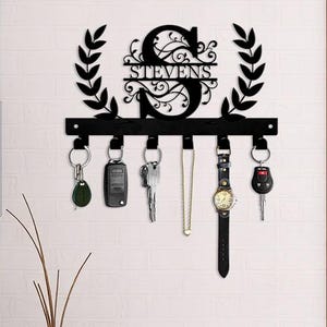 May include: Black metal key holder with the name "STEVENS" and the letter "S" in a decorative font. The key holder has a row of hooks with keys, a car key fob, a watch, and a necklace hanging from them. The key holder is mounted on a white brick wall.