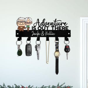 May include: Black metal key holder with the text "Adventure is Out There" and the names "Jack & Kellie". The key holder features cartoon figures and holds keys, a watch, and a necklace. The key holder is mounted on a white wall.