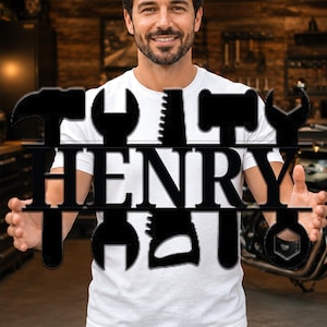 May include: A black metal wall sign featuring the name HENRY in bold capital letters, integrated with silhouettes of various hand tools including a hammer, a wrench, and a saw, designed as a decorative piece for a workshop or garage space.