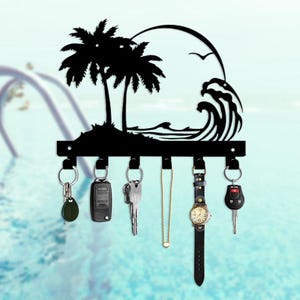 May include: Black metal key holder featuring a tropical beach scene with palm trees, waves, and a sun. The key holder has six hooks, with keys, a watch, and a necklace hanging from them. The background is a light blue.