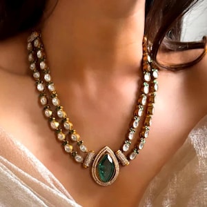 May include: A two-strand gold necklace with clear and green gemstones. The necklace features a large, teardrop-shaped green gemstone pendant. The necklace is worn on a person's neck.