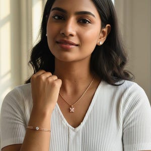 May include: A woman wearing a rose gold butterfly pendant necklace, matching bracelet, and stud earrings. The jewellery is set against a white ribbed top. The butterfly pendant has pink accents.